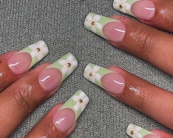 May include: Close-up of a hand with long, light green and white nail polish with white flower accents. Each nail has a small gold dot in the center of the flower.
