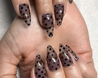 May include: Close-up of a hand with long, pointed nails painted with a sheer, light brown polish and black polka dots.