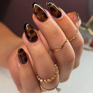 May include: Oval-shaped, tortoise shell-patterned acrylic nails. The nails are a mix of dark brown and amber hues, with a glossy finish. The hand also features several gold rings, including a chain-link band and a chevron ring.