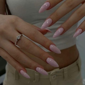 May include: Close-up of hands with long, pointed, light pink acrylic nails. A silver ring with a floral design is on one finger. The nails have a glossy finish. The person is wearing a white top and tan pants.
