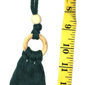 Puede incluir: Un borla de color verde oscuro con un anillo de madera y una cuenta. La borla mide aproximadamente 30 cm de largo.