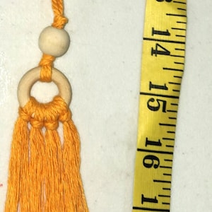 Puede incluir: Una borla naranja con un anillo y una cuenta de madera, junto a una cinta métrica amarilla. La borla mide aproximadamente 45 cm de largo. La borla está hecha de hilo naranja.