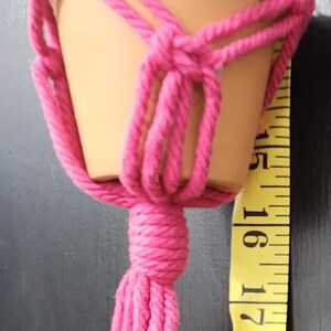 May include: A pink macrame plant hanger with a terracotta pot. The hanger has a tassel at the bottom and is hanging from a yellow measuring tape.