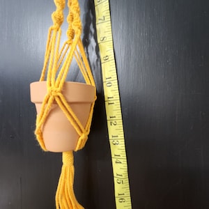 Puede incluir: Un macetero de macramé amarillo con una maceta de terracota. El macetero mide aproximadamente 30 cm de largo.