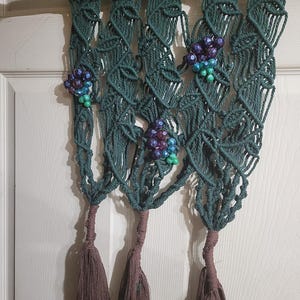 Può includere: Applique da parete in macramè fatto a mano in verde acqua, con un design a vite con grappoli di perline nei toni del viola, blu e verde. La parte inferiore ha nappe marroni. Il macramè è sospeso a un ramo di legno naturale.