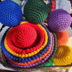 Pode incluir: Uma coleção de itens coloridos de crochê, incluindo esferas e anéis empilhados, em uma variedade de cores como azul, verde, roxo, laranja, amarelo e vermelho. Os itens estão contidos dentro de uma bolsa transparente com zíper.