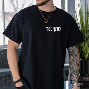 May include: Black short-sleeved t-shirt with the word "TECHNO" printed in white, gothic-style lettering across the chest. The shirt is a solid black color and appears to be made of a soft material.