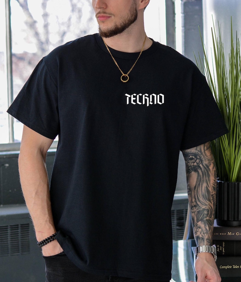 May include: Black short-sleeved t-shirt with the word "TECHNO" printed in white, gothic-style lettering across the chest. The shirt is a solid black color and appears to be made of a soft material.