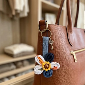 May include: A brown leather tote bag features a hanging keychain accessory. The keychain includes a denim fabric strap, a bronze-toned skeleton key charm, and a fabric flower with petals in white, orange, and dark blue, centered with yellow and brown beads.