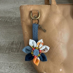 May include: A fabric flower bag charm featuring denim and patterned white petals with orange and brown accents. The center is adorned with small orange and yellow beads surrounding a denim circle. A metal key charm hangs from the denim strap.