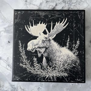 May include: Black and white illustration of a moose with large antlers, surrounded by foliage. The image is printed on a black background with a distressed texture.