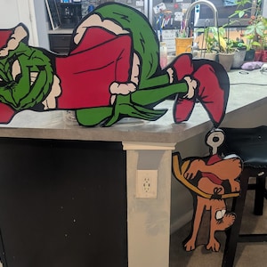 May include: A large, flat, cutout-style wooden decoration featuring a green character wearing a red Santa hat and red shirt, lying down. Attached below is a smaller, separate cutout of a brown dog with a red collar, hanging upside down.