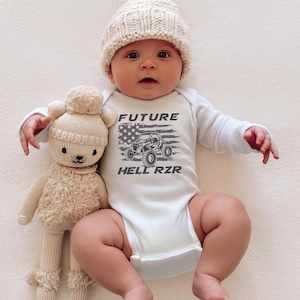 Könnte beinhalten: Ein weißer Baby-Strampler mit langen Ärmeln zeigt eine graue Grafik eines Geländewagens mit einer amerikanischen Flagge und dem Text "FUTURE HELL RZR". Ein Baby trägt den Strampler und hält ein beigefarbenes Stofftier.