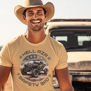 Puede incluir: Una camiseta de color beige con un gráfico de un vehículo todoterreno de camuflaje con el texto "Hell Azar Safety 3rd" en un círculo alrededor.