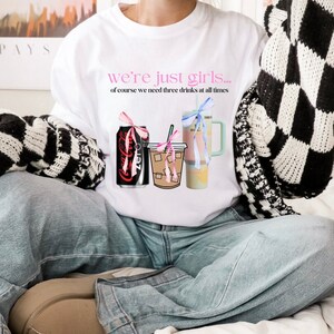 Könnte beinhalten: Weißes Sweatshirt mit dem rosa Text "we're just girls... of course we need three drinks at all times". Die Grafik zeigt eine Limonadendose, Eiskaffee und einen Becher, jeweils mit einer Schleife. Die Person trägt eine blaue Jeans.