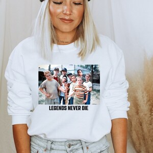 May include: White sweatshirt featuring a graphic of a group of boys. The graphic includes the text "LEGENDS NEVER DIE" in black block letters. The boys are wearing various shirts and hats.