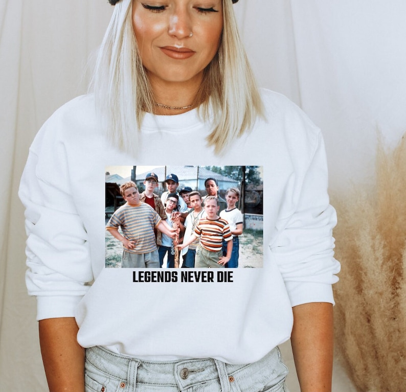 May include: White sweatshirt featuring a graphic of a group of boys. The graphic includes the text "LEGENDS NEVER DIE" in black block letters. The boys are wearing various shirts and hats.