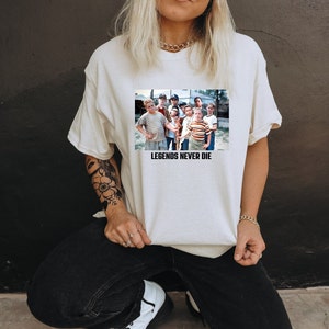 May include: Cream-colored t-shirt featuring a rectangular graphic of a group of boys. The graphic is a still from a movie. The text "LEGENDS NEVER DIE" is printed below the graphic in black. The shirt is made of a soft material.