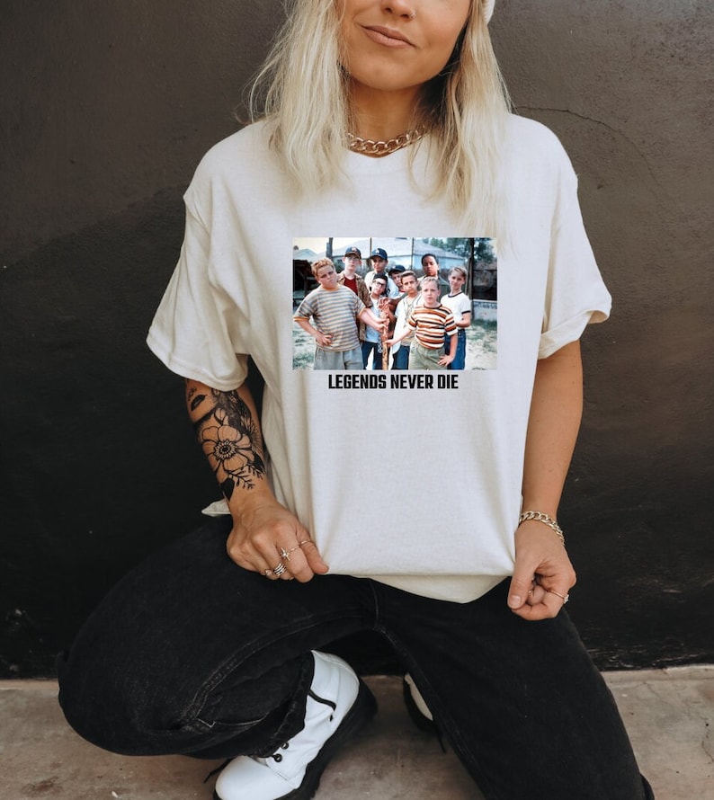 May include: Cream-colored t-shirt featuring a rectangular graphic of a group of boys. The graphic is a still from a movie. The text "LEGENDS NEVER DIE" is printed below the graphic in black. The shirt is made of a soft material.
