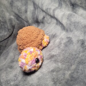 May include: A small, colourful, crocheted turtle toy with a brown shell and a pink, yellow, and orange patterned body. The turtle has black eyes and a pink nose.