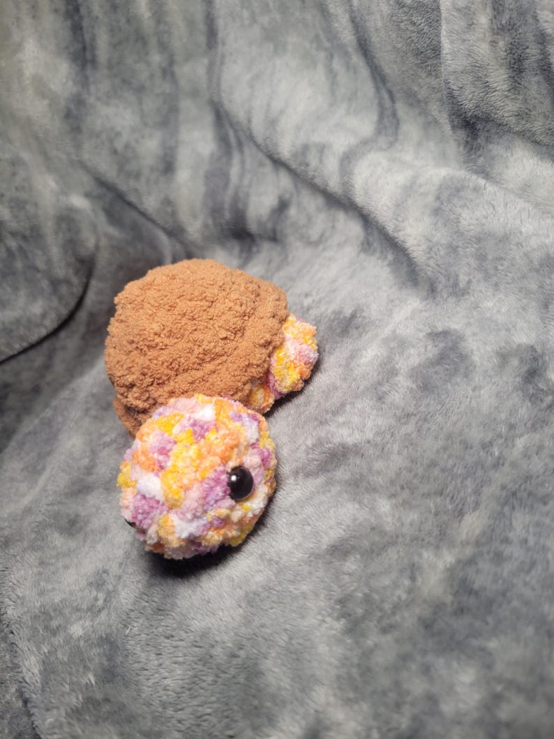 May include: A small, colourful, crocheted turtle toy with a brown shell and a pink, yellow, and orange patterned body. The turtle has black eyes and a pink nose.