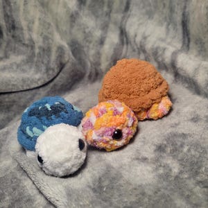 May include: Three crocheted turtle plushies. One is blue and white, one is orange, pink, and white, and one is brown.