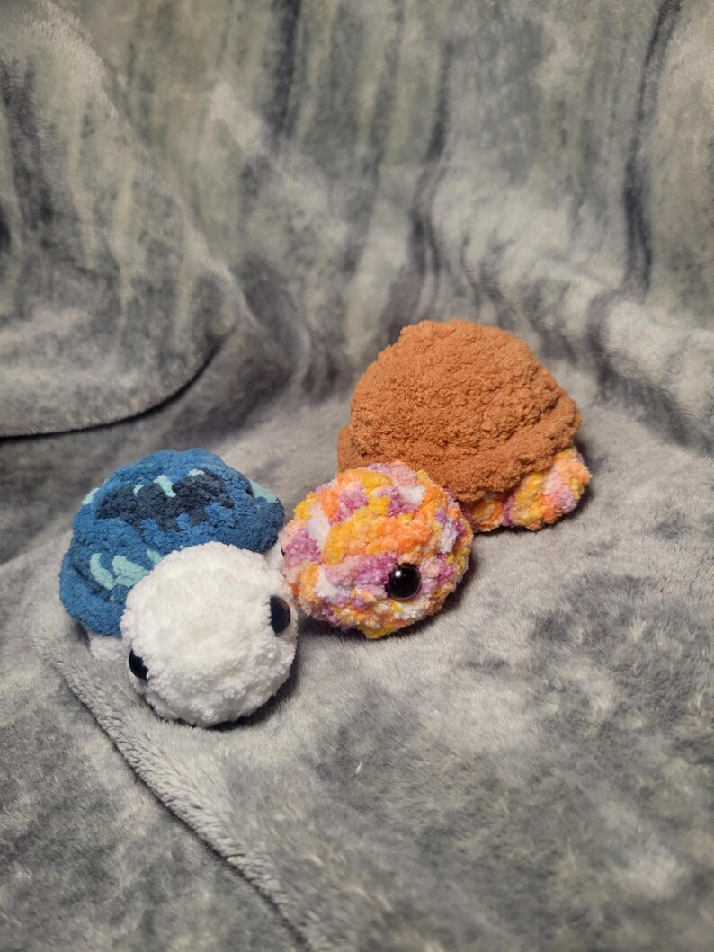 May include: Three crocheted turtle plushies. One is blue and white, one is orange, pink, and white, and one is brown.