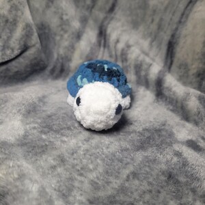 May include: A small crocheted turtle toy with a blue and white shell and white face. The toy has black eyes and a white body.