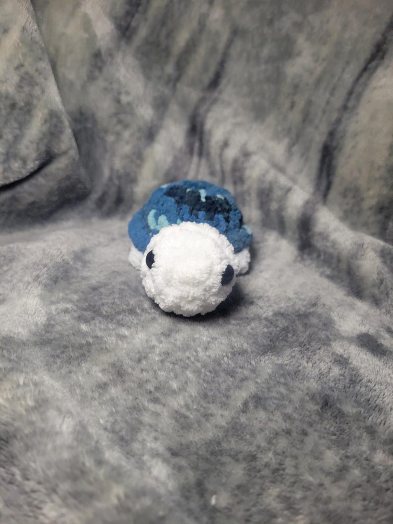 May include: A small crocheted turtle toy with a blue and white shell and white face. The toy has black eyes and a white body.