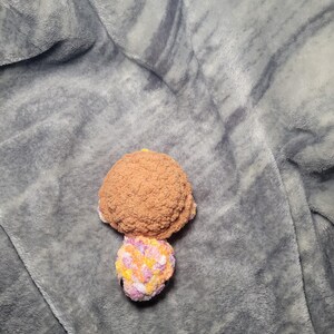 May include: A crocheted stuffed turtle toy with a brown shell and a multi-coloured head and legs. The toy is lying on a grey fuzzy blanket.