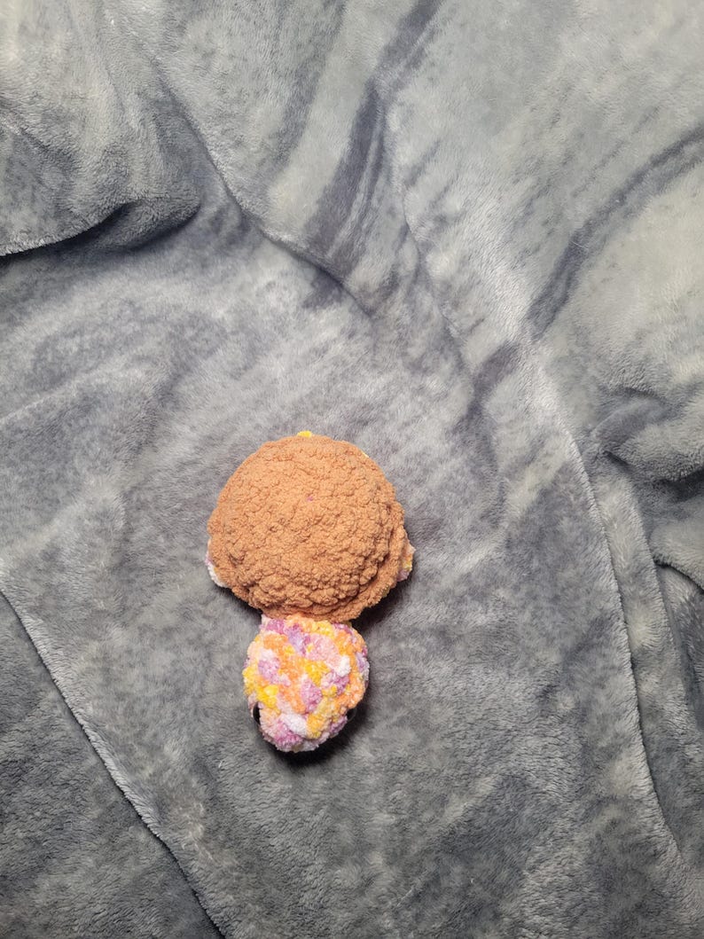 May include: A crocheted stuffed turtle toy with a brown shell and a multi-coloured head and legs. The toy is lying on a grey fuzzy blanket.