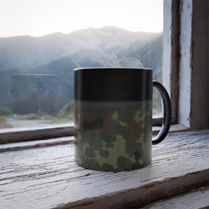 May include: A black ceramic mug with a camouflage pattern that appears when hot liquid is added. The mug is sitting on a weathered wooden surface.