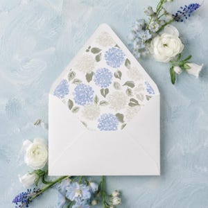 May include: A white envelope with an open triangular flap featuring an interior liner pattern of watercolor-style blue and white hydrangea blossoms with green leaves, surrounded by fresh white ranunculus and blue delphinium flowers.