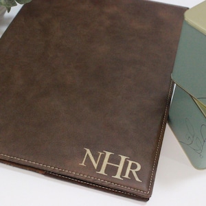 May include: A brown leather portfolio with the initials "NHR" embossed in a lighter color. The portfolio is rectangular and has a stitched border. A decorative tin with a gold lid and floral design is next to the portfolio.