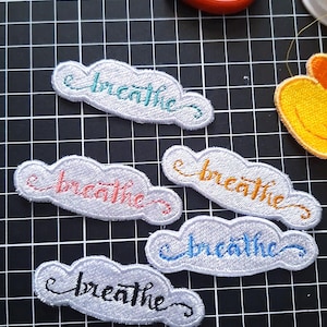 May include: Five embroidered cloud-shaped patches, each with the word "breathe" in a different colour: teal, coral, orange, blue, and black. The patches are white with a grey border, suitable for clothing or accessories.