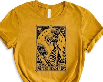 May include: A mustard yellow t-shirt with a black and white illustration of a skeleton reading a book. The illustration is framed in a rectangle with the text "THE READER" at the bottom.