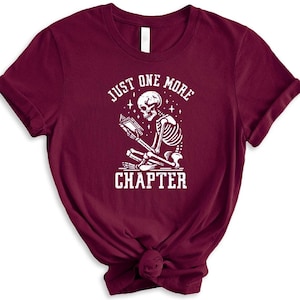 May include: A maroon t-shirt featuring a white graphic of a skeleton reading a book. The graphic reads "Just One More Chapter".