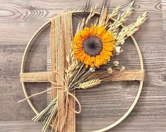 May include: A circular wall hanging featuring a bright yellow sunflower and wheat stalks. The floral arrangement is secured to a wooden hoop with natural twine. The background is a wooden surface.