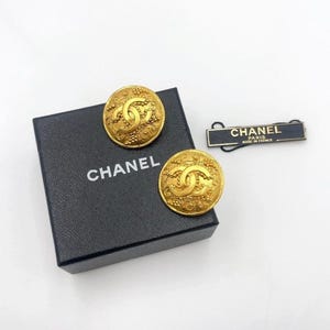 May include: A pair of gold-toned Chanel earrings with a textured design and the iconic interlocking CC logo. The earrings are round and sit on a black box with the brand name in white. A black tag with gold text reads "CHANEL PARIS MADE IN FRANCE."