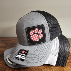 May include: Gray and black trucker hat with a red and white checkered paw print patch. The hat has a black mesh back and a gray front with the text "Richardson. Original 112 Trucker Adjustable" on the front.