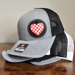 May include: A gray and black trucker hat with a red and white checkered heart patch. The hat has a black mesh back and a gray front with a black brim. The patch is sewn onto the front of the hat.