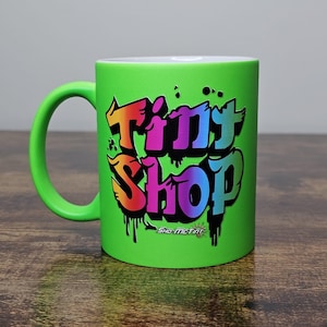 May include: A green ceramic mug with a colorful graffiti-style design that says "Tiny Shop" with the words "Sho-Me-Tiny" underneath.