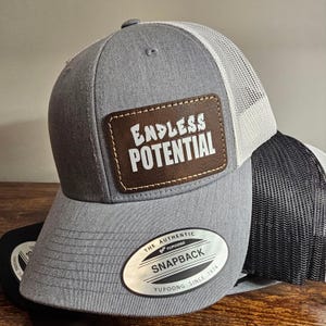 May include: Gray and white trucker hat with a brown leather patch that says "Endless Potential". The hat has a white mesh back and a YUPOONG logo on the side.