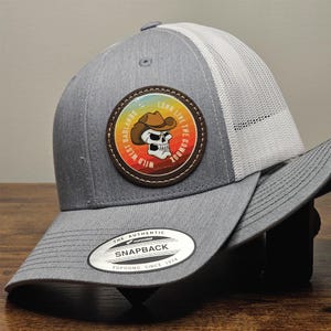 May include: Gray and white trucker hat with a brown leather patch featuring a skull wearing a cowboy hat and the text "Wild West Badlands Long Line The Cowboy". The hat has a "The Authentic Yupoong Snapback Yupoong Since 1974" label.