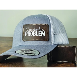 May include: Gray and white trucker hat with a brown leather patch that says "Somebody's Problem" in white cursive lettering. The hat has a brown leather strap and a silver metal tag that says "The Authentic Snapback Yupoong Since 1974".