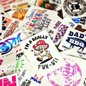 May include: Assortment of colorful temporary tattoos with various designs, including a mushroom character with the text "I'm a really fun-gi!" and a BBQ design with the text "Dad's World Famous BBQ Chillin' & Grillin' King".