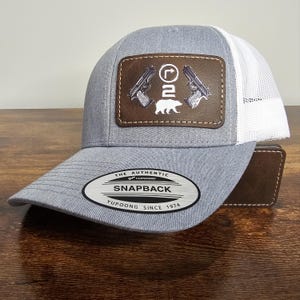 May include: A gray snapback hat with a white mesh back. The front features a brown leather patch with two pistols, the number 2, and a bear silhouette. The hat has a curved brim and the text "SNAPBACK" and "YUPOONG SINCE 1974".