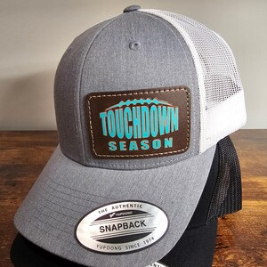 May include: A gray and white trucker hat with a brown leather patch that says "TOUCHDOWN SEASON" in turquoise and orange lettering. The hat has a black mesh back and a YUPOONG label.