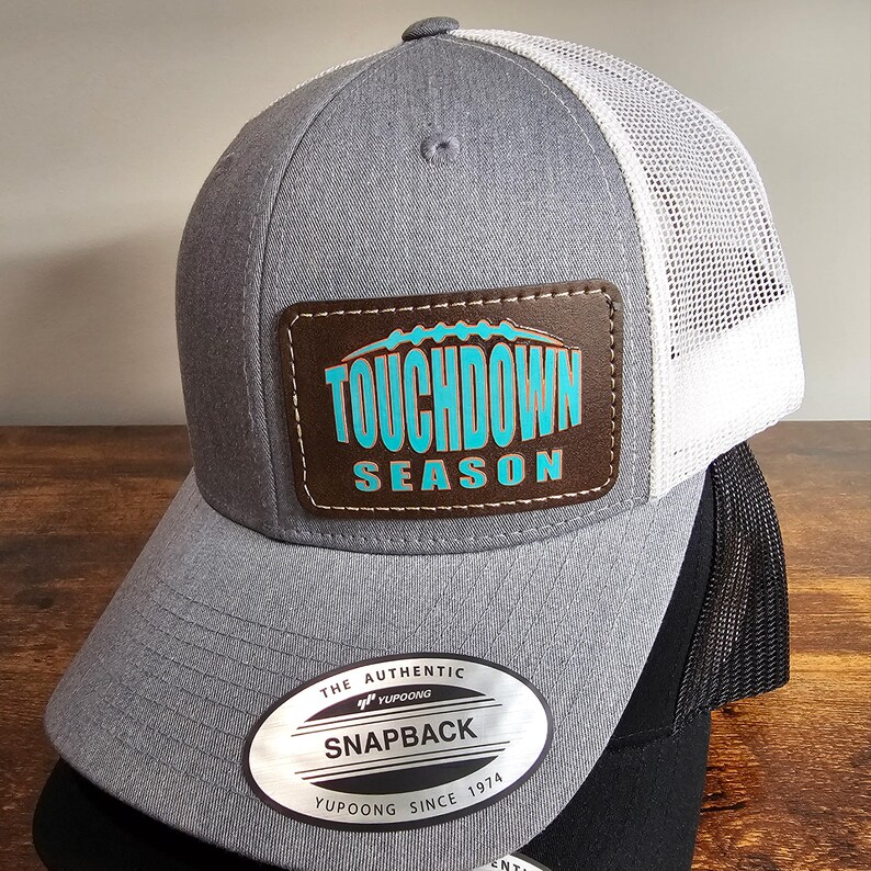 May include: A gray and white trucker hat with a brown leather patch that says "TOUCHDOWN SEASON" in turquoise and orange lettering. The hat has a black mesh back and a YUPOONG label.