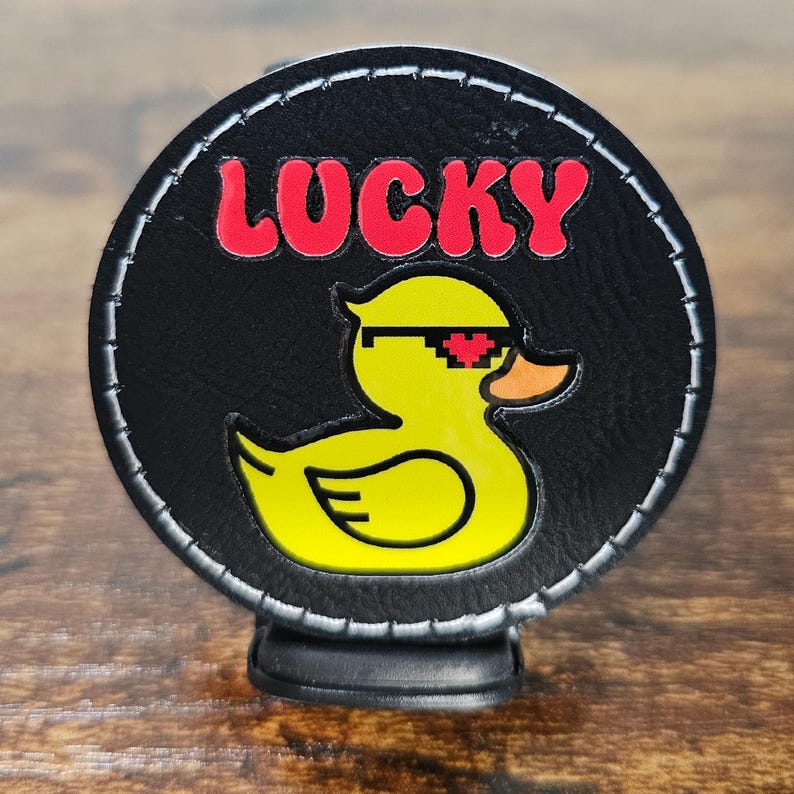 May include: A black round patch with a yellow rubber duck wearing sunglasses and a red heart over one eye. The text "LUCKY" is written in red above the duck.